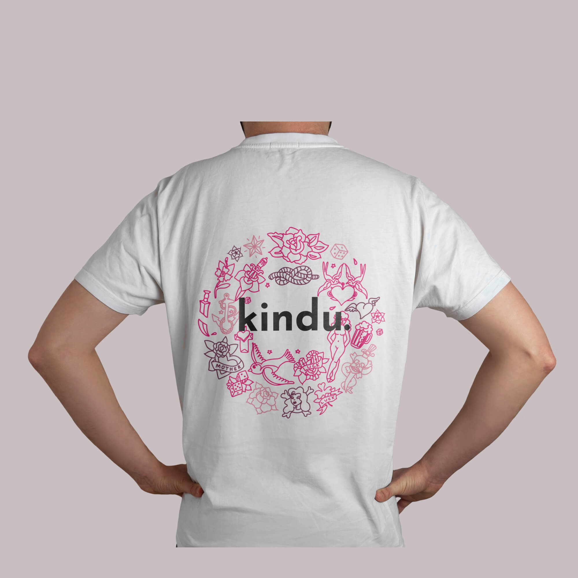 Imagen de la camiseta blanca por detrás de la tienda de Kindu, rotulada con el logo en el centro rodeado de dibujos contoneados en tonos rosas y malvas con un estilo tattoo.
